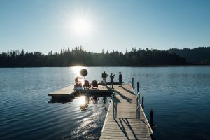 A group of people gathered on a dock by the water, surrounded by trees, with boats nearby and a scenic sunset in the background.