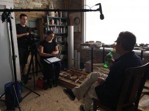 A group of people gathered in a room filled with furniture, including a table and bookcase, surrounded by walls and a floor. Some individuals are reading books, and they are wearing various clothing and footwear styles.