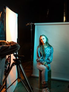 A young woman illuminated in blue light is seated in a photo studio, posing for a photograph.