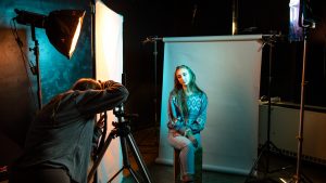 A young woman illuminated in blue light is seated in a photo studio, posing for a photograph.