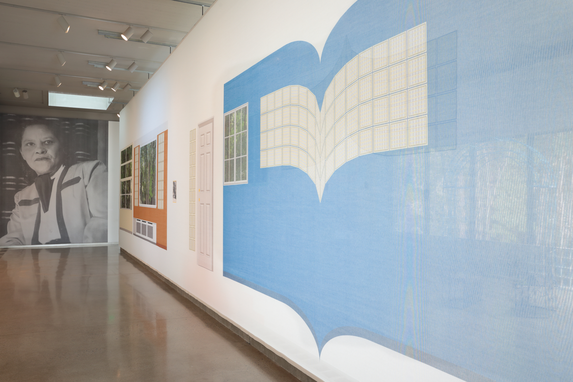 A wide shot of the exhibiton Dear Mazie, showing a large scale black and white photo of her taking up the entire back wall and a translucent fabric on the right printed with some architecural drawings by Amaza Lee Meredith and acting as a divider in the space.