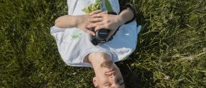 A man laying in grass in the sunshine, eyes closed and clutching a camera to his chest