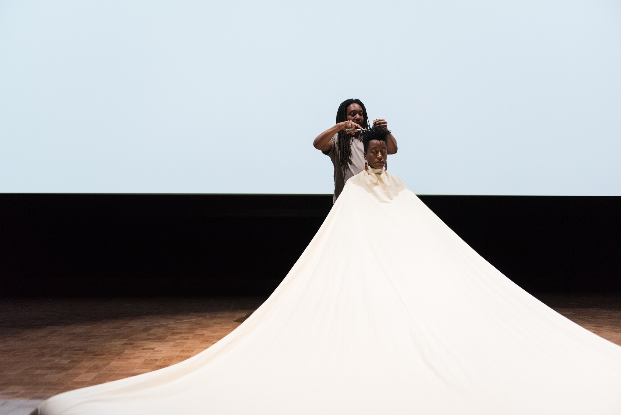 person dancing under a large, ballooning white fabric cloth white a person cuts their hair behind them