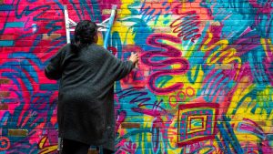 A woman on a ladder spray paints a colorful abstract mural on a brick wall