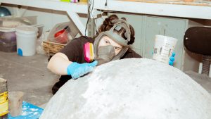 A student wearing protective breathing equipment builds a large round sculpture in their studio