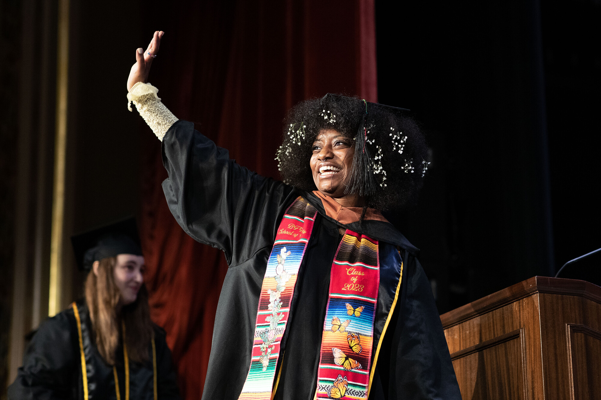 A smiling graduating student in regalia waves to the audience