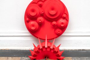 A large red sculpture, the top portion is a circle with raised circles inside of it, the botton is a semi cirlce rigned with spikes, resting on a white wall and sitting on a brown checkerboard floor