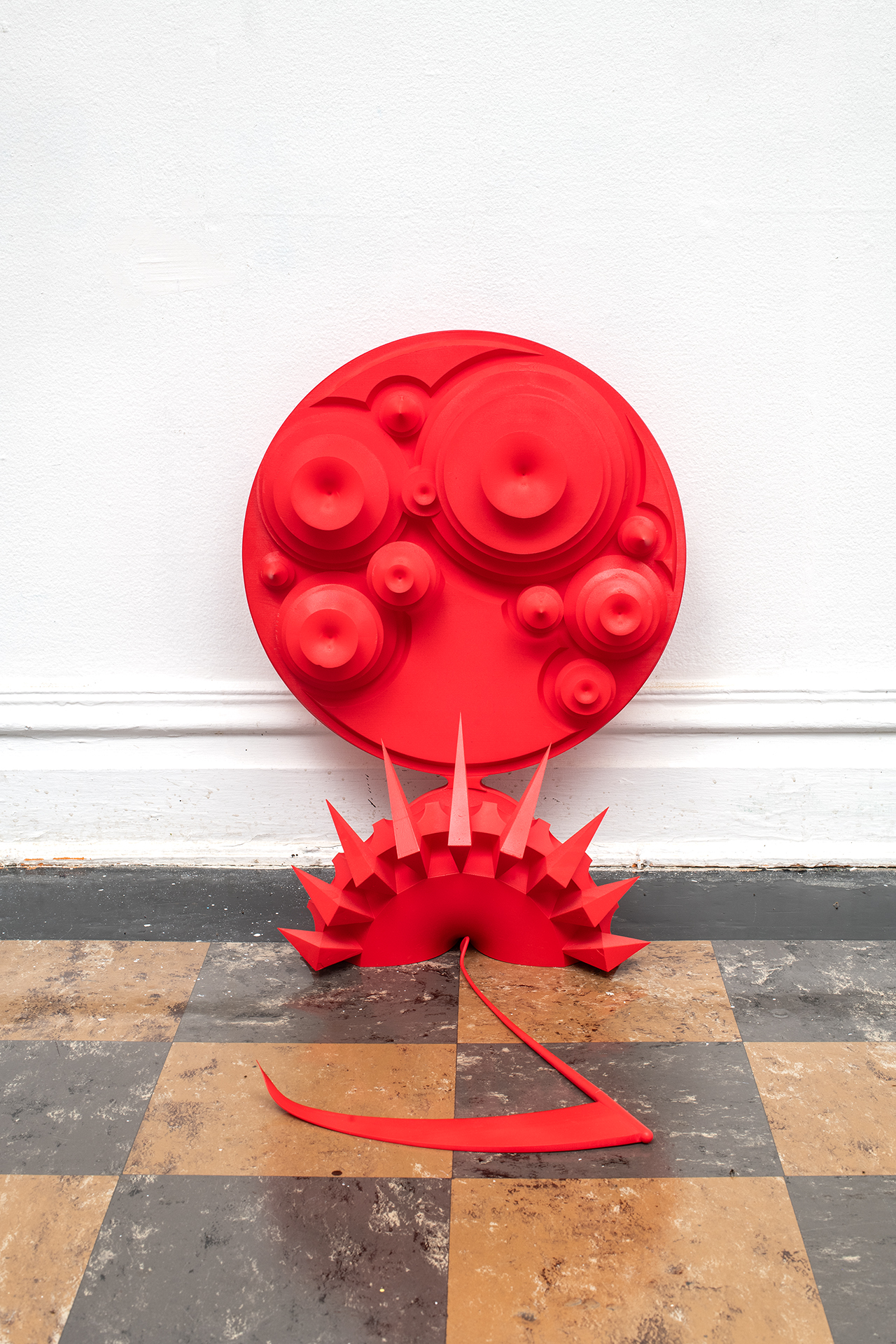 A large red sculpture, the top portion is a circle with raised circles inside of it, the botton is a semi cirlce rigned with spikes, resting on a white wall and sitting on a brown checkerboard floor