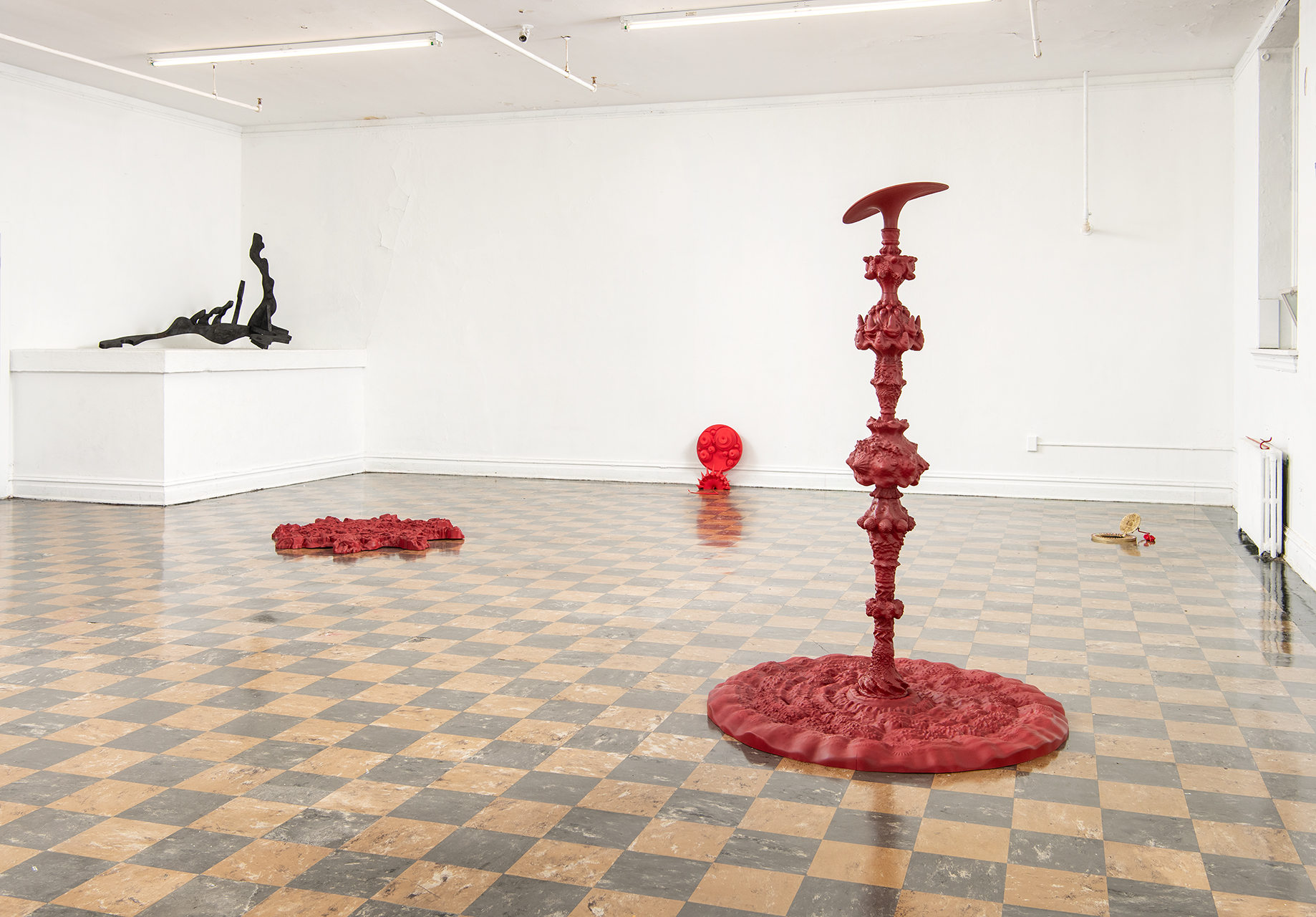 Exhibition view of Zhao's work, showing 4 red sculpture's of varying sizes on a brown checkerboard floor, as well as the black sculpture resting on a white box higher up.