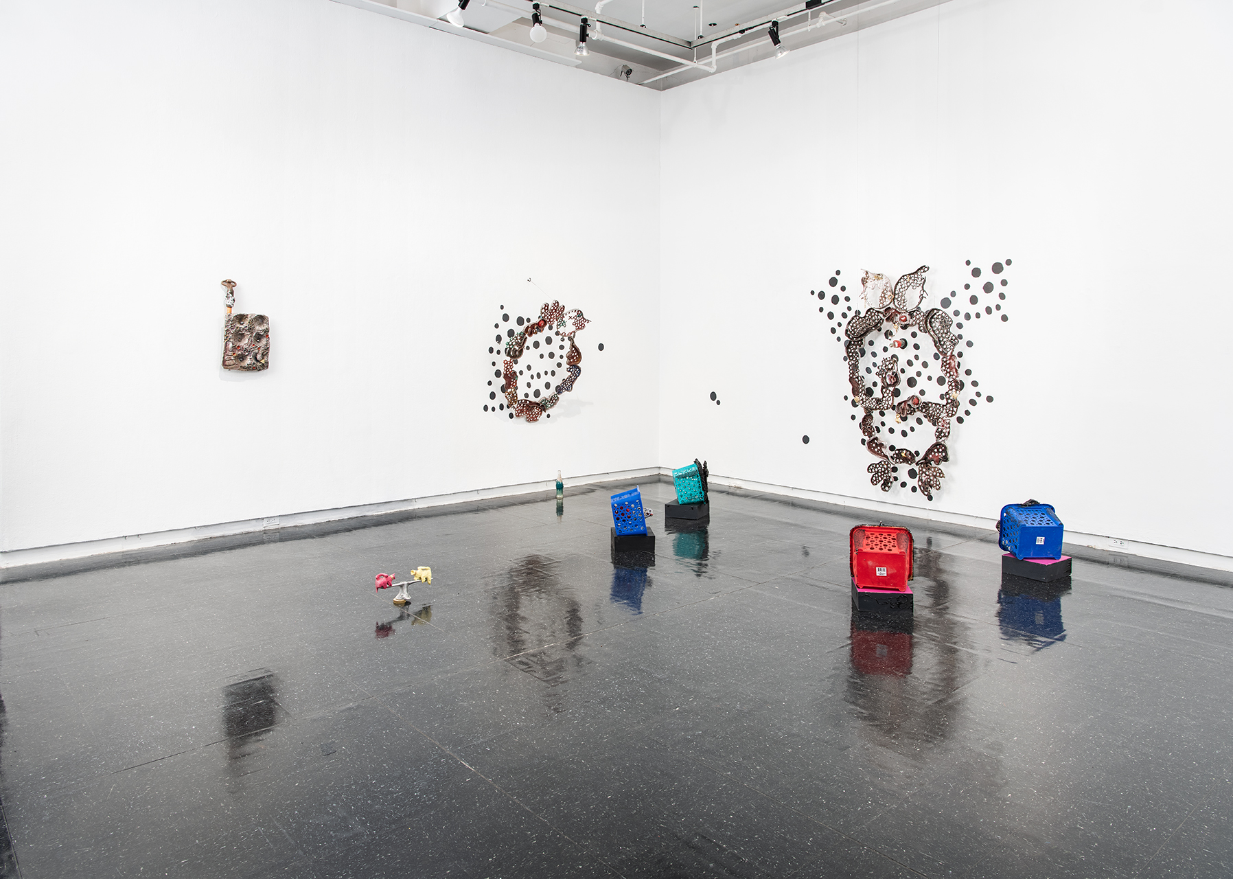 Exhibition view of Mupita's full installation showing 3 sculptures mounted on the wall varying in scale with black dots, metal organic forms, and peepholes, as well as floor sculptures made from plastic crates.