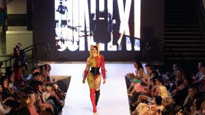 model walking down the center of a runway dressed in a punk red black and leopard print student designed outfit with spiky hair and goth inspired makeup with the word CONTEXT on a large screen behind them