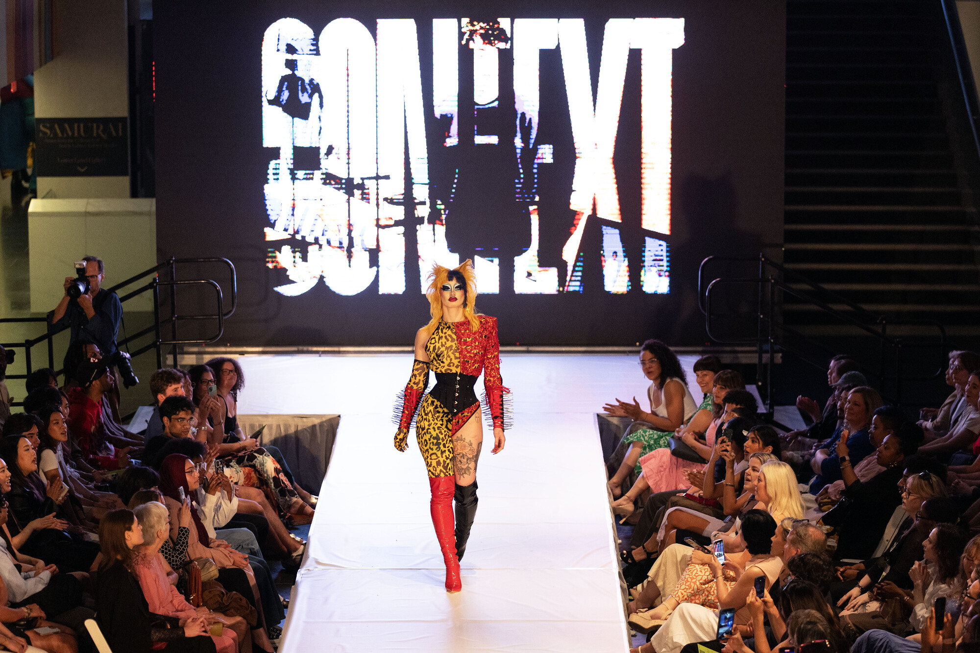 model walking down the center of a runway dressed in a punk red black and leopard print student designed outfit with spiky hair and goth inspired makeup with the word CONTEXT on a large screen behind them