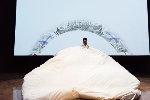 person dancing under a large, ballooning white fabric cloth white a video displaying a grey and blue abstract gradient pattern plays behind them