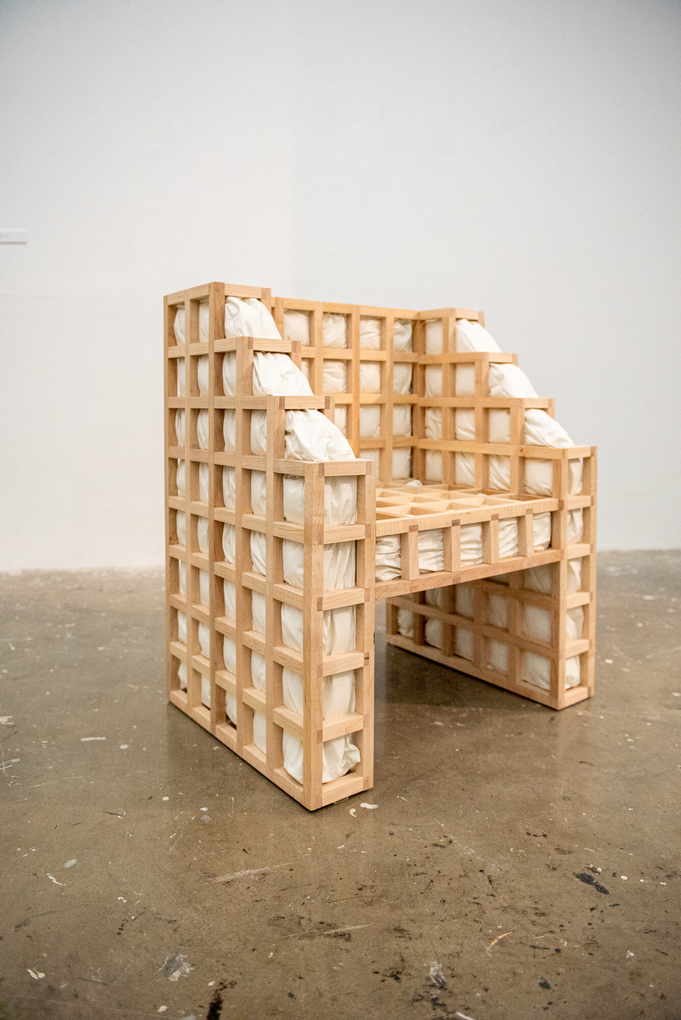 Secrets: squished is a functional chair with a wooden grid exoskeleton and protruding white cushions. The bulging cushions interrupt the architectural grid structure. Except for upholstery foam and batting, the contents of the cushions are known only to the artist.