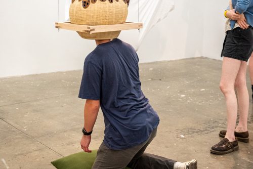 Circular ceramic piece hovering above the floor with a person kneeling on the floor and placing their head into the ceramic piece like a hat