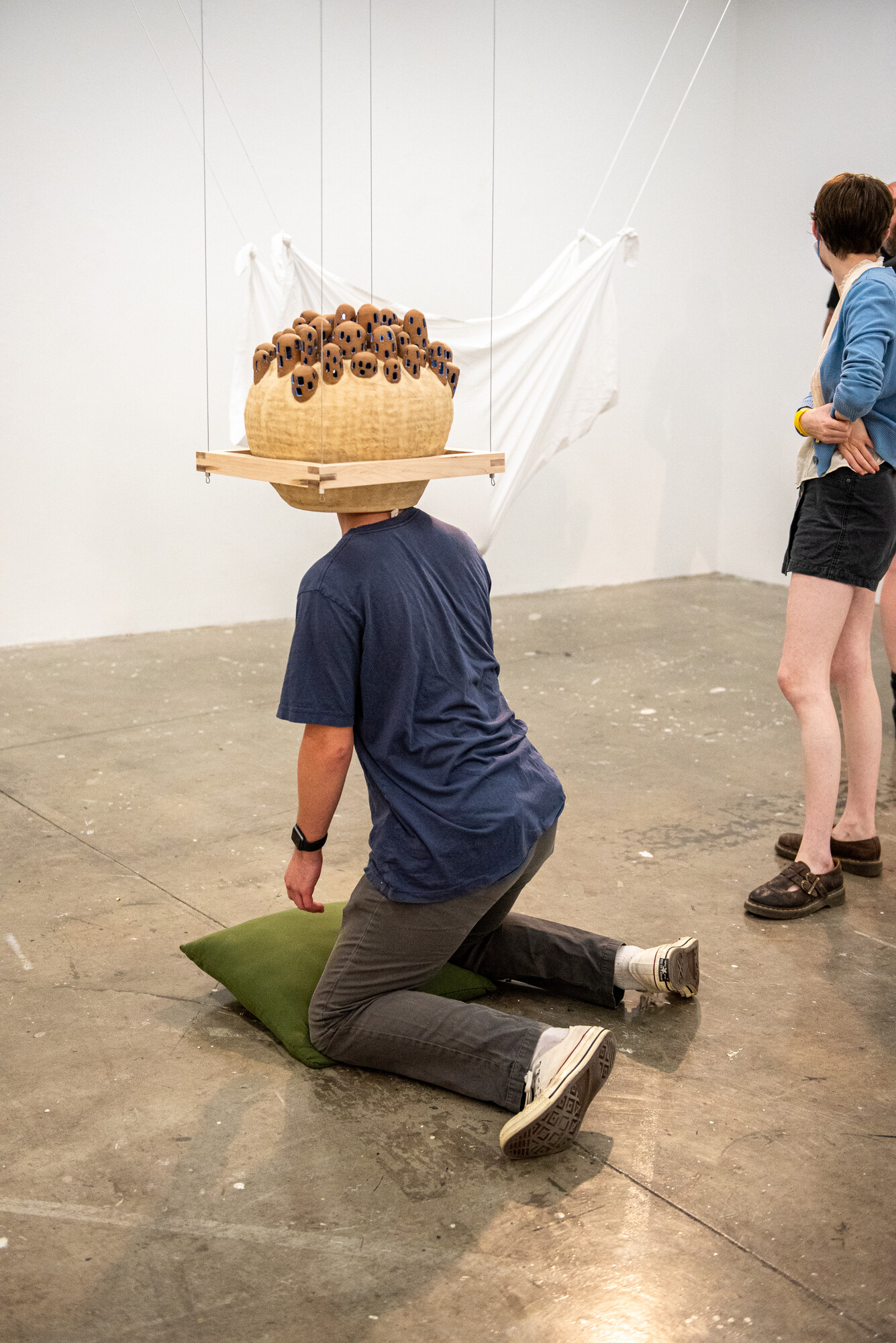 Circular ceramic piece hovering above the floor with a person kneeling on the floor and placing their head into the ceramic piece like a hat