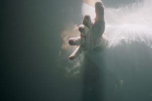 Viewing from underwater, there is a person floating just below the surface of the water. The face is obscured as a pruney hands is outstretched to the viewer.