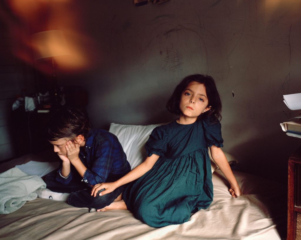 A woman sitting on a bed with a young girl on her lap in a cozy bedroom setting. The bed is covered with soft bedclothes, and the walls are simple and comforting.