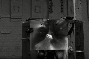 A black and white photo shows a large hollow kiln on the bottom right side of the image. There are two people hiding inside the kiln they are covering their faces and bodies with a large reflective piece of glass that is magnifying their faces. 