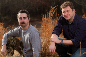 A couple of men smiling in a grassy field, wearing casual clothing, surrounded by straw and plants.