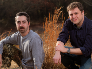 A couple of men smiling in a grassy field, wearing casual clothing, surrounded by straw and plants.