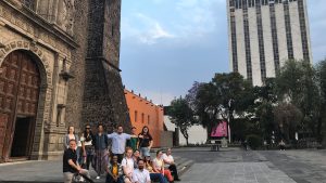 A group of students and faculty stand and sit on the steps in front of an old cathedral in Mexico. They are all smiling and looking at the camera.