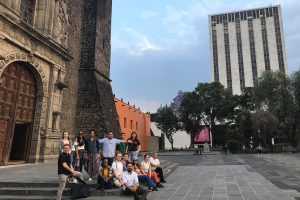 A group of students and faculty stand and sit on the steps in front of an old cathedral in Mexico. They are all smiling and looking at the camera.