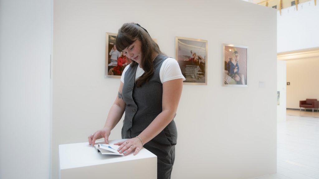 A woman standing at a desk looking through an art book in an indoor art gallery, surrounded by walls adorned with artwork. 