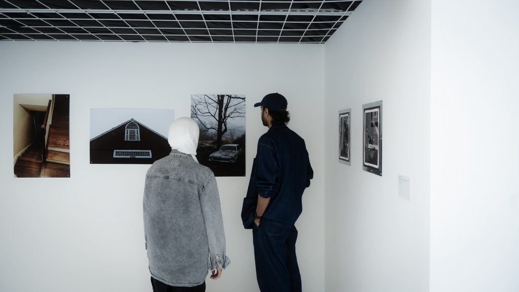 A man and a woman stand next to each other in an art gallery, looking at a painting on the wall. The man is dressed in a coat, and they are surrounded by various artworks displayed in picture frames. The exhibition takes place indoors.