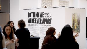 A group of people standing in front of a white gallery wall displaying the text "TO THINK WE WERE EVER APART" accompanied by the date "09.24-10.30.24". The setting is an indoor art gallery or exhibition room, featuring various individuals, including men and women dressed in casual clothing.