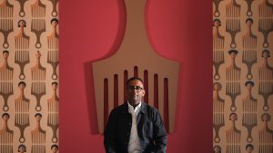 Jomo is sitting on a chair in the shape of a hair pic. Behind him is an installation of another artwork of his: a large wooden hair pic on hung on the wall. The wall has wallpaper with illustrations of hair pics.