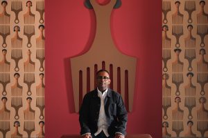 Jomo is sitting on a chair in the shape of a hair pic. Behind him is an installation of another artwork of his: a large wooden hair pic on hung on the wall. The wall has wallpaper with illustrations of hair pics.