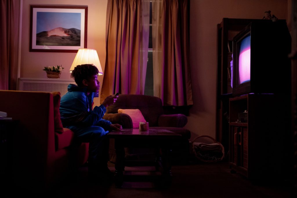 A man sitting in a chair in a living room, surrounded by furniture and curtains, with a television visible. There is also a child nearby, and the room features a window and floor.