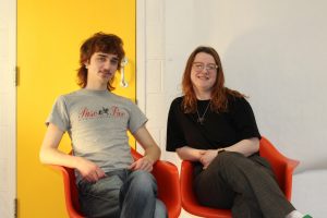 two people sitting in red chairs beside each other in front of a white wall