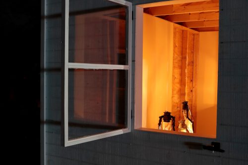The side of the white shed has an open window. Inside, there are three gas lamps, glowing the interior of the shed.