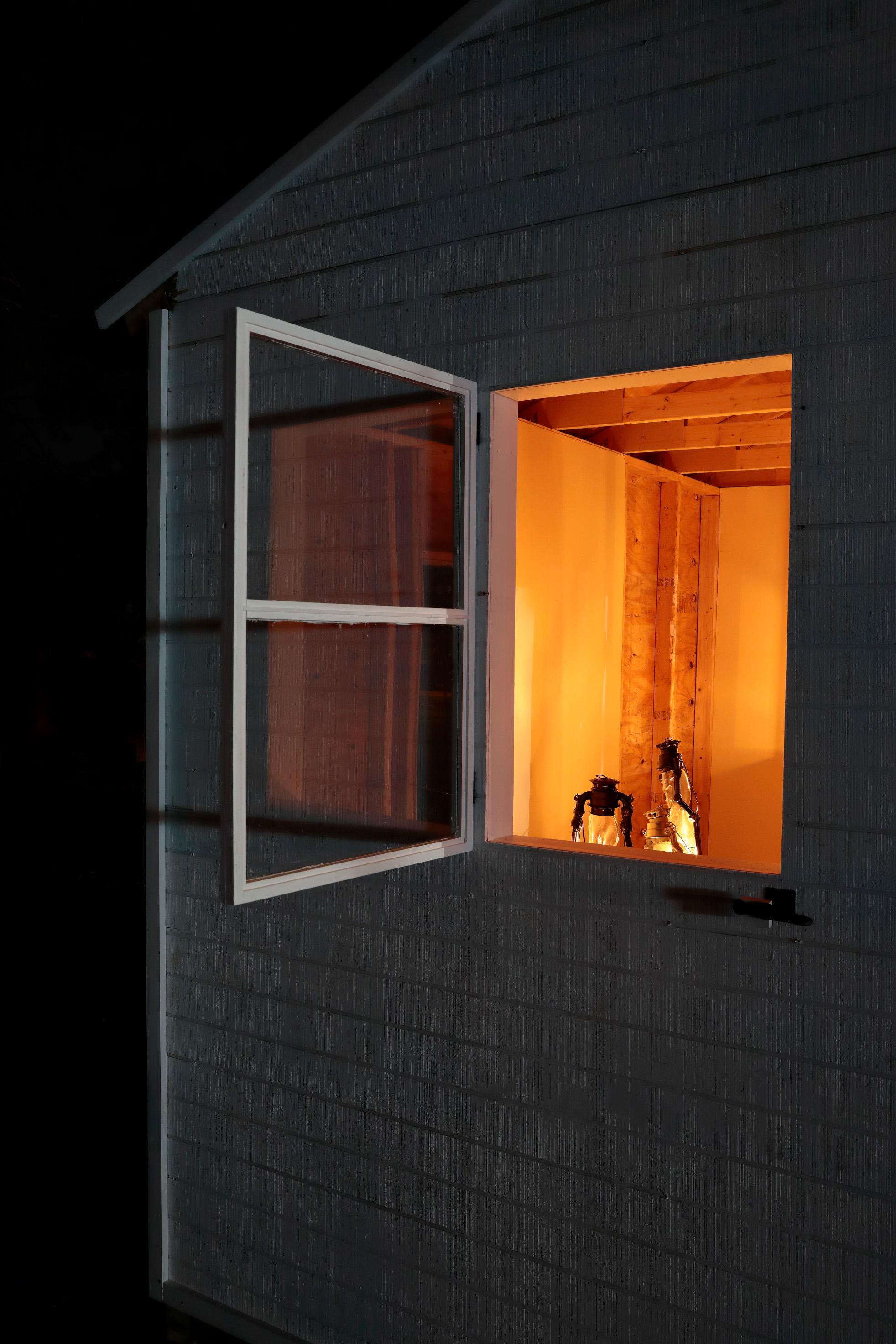 The side of the white shed has an open window. Inside, there are three gas lamps, glowing the interior of the shed.