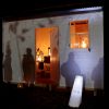 A white shed is illuminated in the dark. Inside are old gas lamps glowing with warm yellow light. On the exterior of the shed are shadows of people.