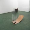 Two sculptures are on a green carpet. The sculpture closest to the viewer has a metal rack is standing upright and attached to a wide and curved piece of wood by a metal and fabric cord. The other sculpture and features a metal rack slight curved and sloping down to the carpet. An "L" shaped piece of wood is attached to the top of the metal rack.