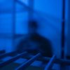 A man with a blurred out face stands in a room lit in blue light.