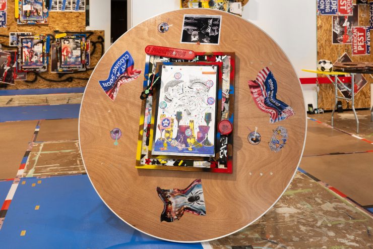 A close-up photo of a larger installation. In the center of the image is a circular wooden table on its side. On the surface of the table top is a painting of a still life with two cartoon figures crying. The frame of the painting is made out of found objects like pieces of chalk, scraps of paper, and a cigarette. Around the painting on the table are crumpled up images of promotional banners with the American flag iconography.