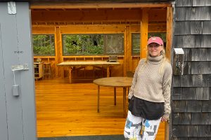 Michelle is leaning against a doorframe smiling. Both the exterior and interior of the building are completely made out of wood.