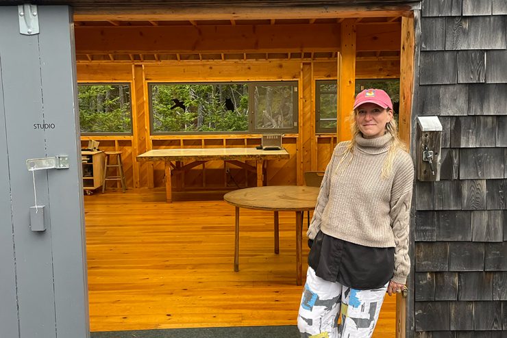 Michelle is leaning against a doorframe smiling. Both the exterior and interior of the building are completely made out of wood.