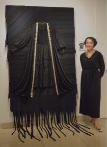 Kim Guthrie, dressed in all black, poses next to her artwork Dead Dress, a black lace dress mounted to a backdrop of black fabric strips