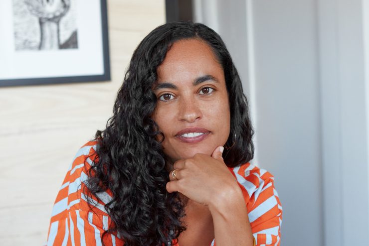 Chloe is leaning on a table, with her elbows resting on the table. One hand is propping up her head. Chloe is smiling and looking at the camera.