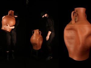 There are three large terracotta vases lined up from left to right in front of a black ground. There is one person in black clothing holding up the left-most vase, and there is a person dressed in black clothing standing next to the middle vase, looking down at it. The right-most vase is the same height as the person standing in the middle.