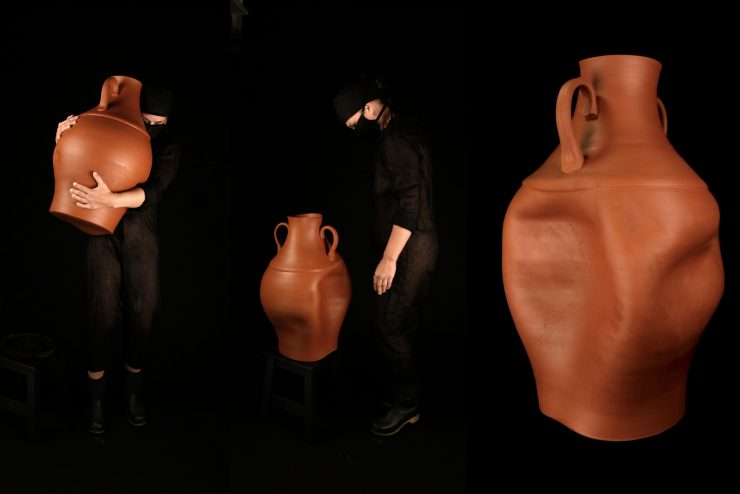 There are three large terracotta vases lined up from left to right in front of a black ground. There is one person in black clothing holding up the left-most vase, and there is a person dressed in black clothing standing next to the middle vase, looking down at it. The right-most vase is the same height as the person standing in the middle.