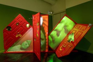 A sculpture consisting of three open red doors in a line. The left and right doors are tilted on their sides. In the door frame are up-close images of a person with green skin with a sad expression. On the doors are different objects, such as a hat, a doorknob made to look like the nose of a sad cartoon face, and flowers.