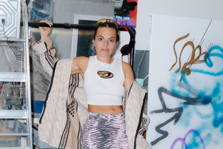 Anna is leaning against a wall, holding onto a pipe that is slightly above her head. Behind her are fragments of her artwork and storage.