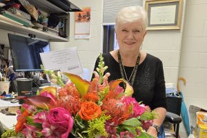 Linda Johnston is wearing a black shirt and necklace, seated at a desk with a bouquet of pink flowers in front of her