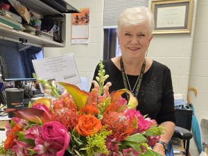Linda Johnston is wearing a black shirt and necklace, seated at a desk with a bouquet of pink flowers in front of her
